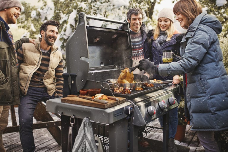 En grupp människor grillar utomhus på vintern på en grill. På grillen ligger kött, grönsaker och en grillad kyckling.
