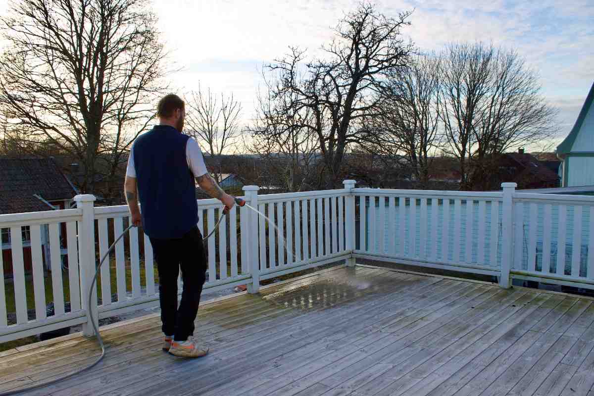 Man som rengör en träterrass med en trädgårdsslang, vitt träräcke, trädgård i bakgrunden.