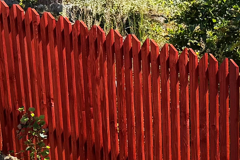 Rött trästaket med spetsiga toppar i en ljus trädgård.