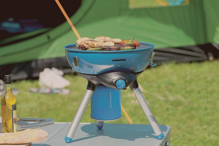 Campingbord med bärbar gasolgrill där kött och grönsaker grillas, med ett tält i bakgrunden