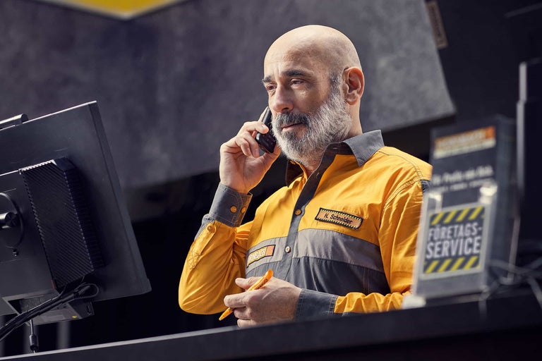 En Hornbach-medarbetare i arbetskläder pratar i telefon vid en servicedisk och tittar på en bildskärm.