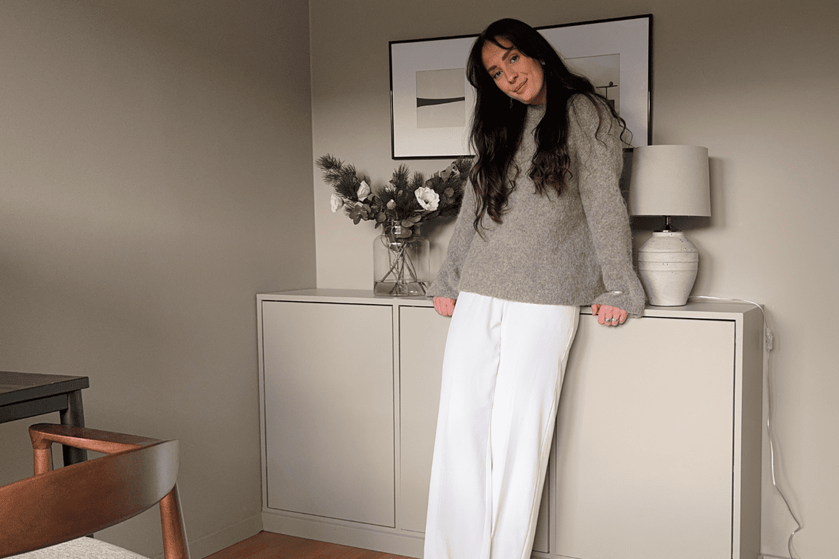 Interiörbild av ett rum med en kvinna som lutar sig mot en byrå, med en tavla ovanför och en vas med blommor på den.