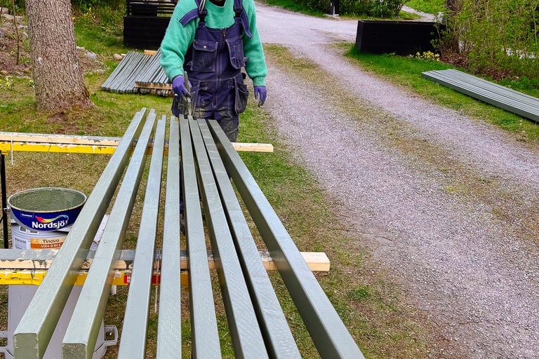 En person målar träribbor med färg utomhus. En färgbehållare av märket Nordsjö syns.