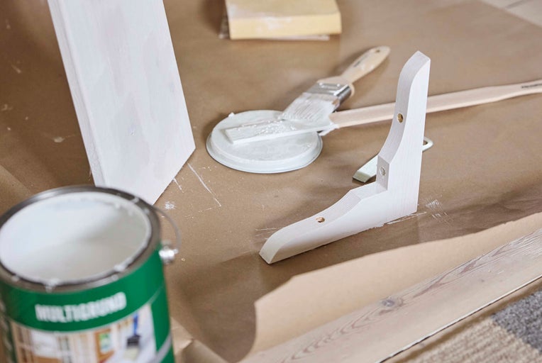 En öppnad burk med grundfärg, en pensel samt vitmålade hyllkonsoler och en träbräda på brunt skyddspapper.