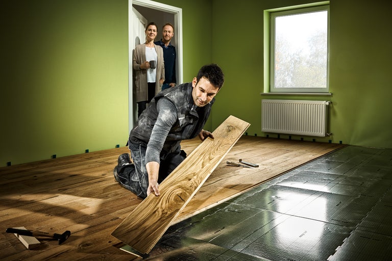 En hantverkare lägger trägolv i ett rum. I bakgrunden står två personer i en dörröppning.