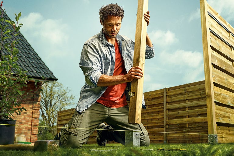 En man monterar ett trästaket i trädgården med trästolpar och stolpfästen