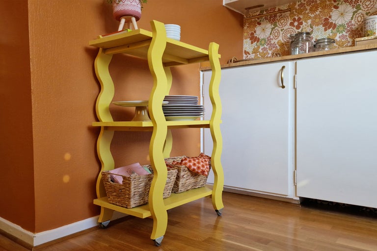 Gul rullvagn med tre nivåer och diverse föremål i ett kök.