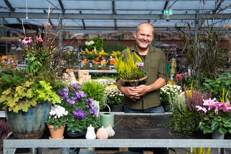 En man håller en blomkruka i ett trädgårdscenter, omgiven av olika växter och blommor.