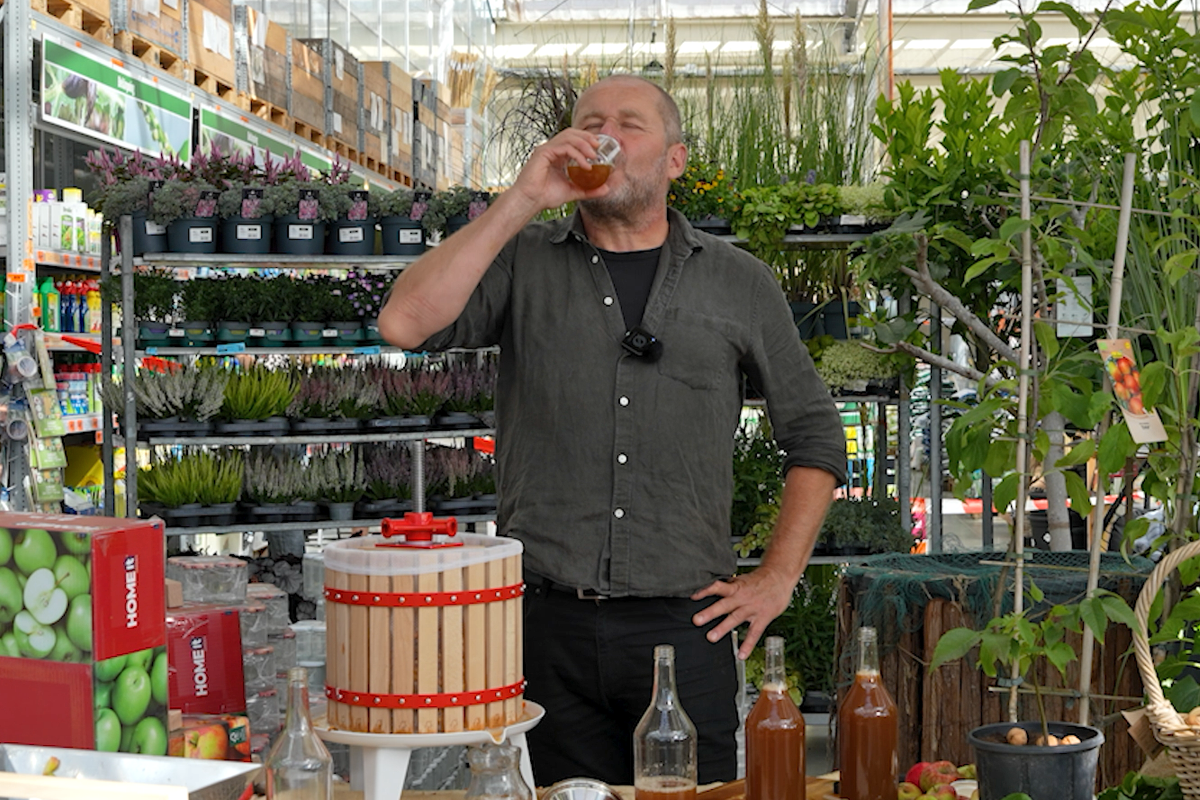 Man dricker färsk äppeljuice vid en fruktpress i trä i en Hornbach butik bland växter och trädgårdstillbehör