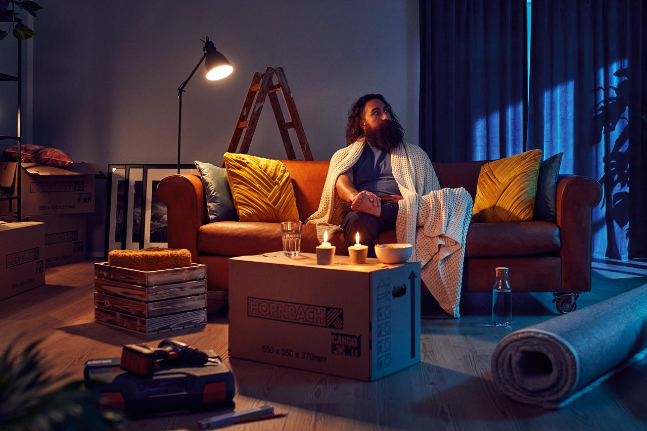 En skäggig man sitter i en soffa i ett vardagsrum under en flytt, omgiven av Hornbach flyttkartonger, ljus och en verktygslåda.
