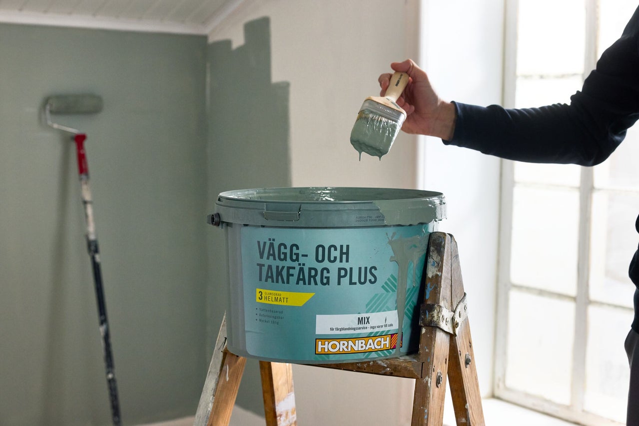 En person håller en pensel över en öppen hink Hornbach vägg- och takfärg på en trappstege av trä i ett rum som målas.
