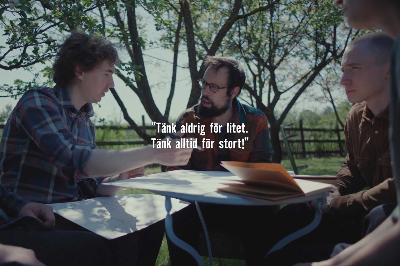 Tre män sitter vid ett bord utomhus under träd och diskuterar planeringsdokument för ett projekt.