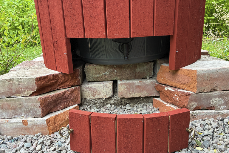 Röd träpanel för regntunna med avtappningskran, stående på en tegelgrund i en trädgård.