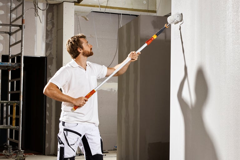 En man målar en vägg med en färgroller på ett teleskopskaft i ett oavslutat rum.