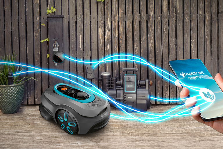 Gardena smart system med robotgräsklippare, bevattningsstyrning och pump, styrt via en smartphone app framför ett trästaket i trädgården.