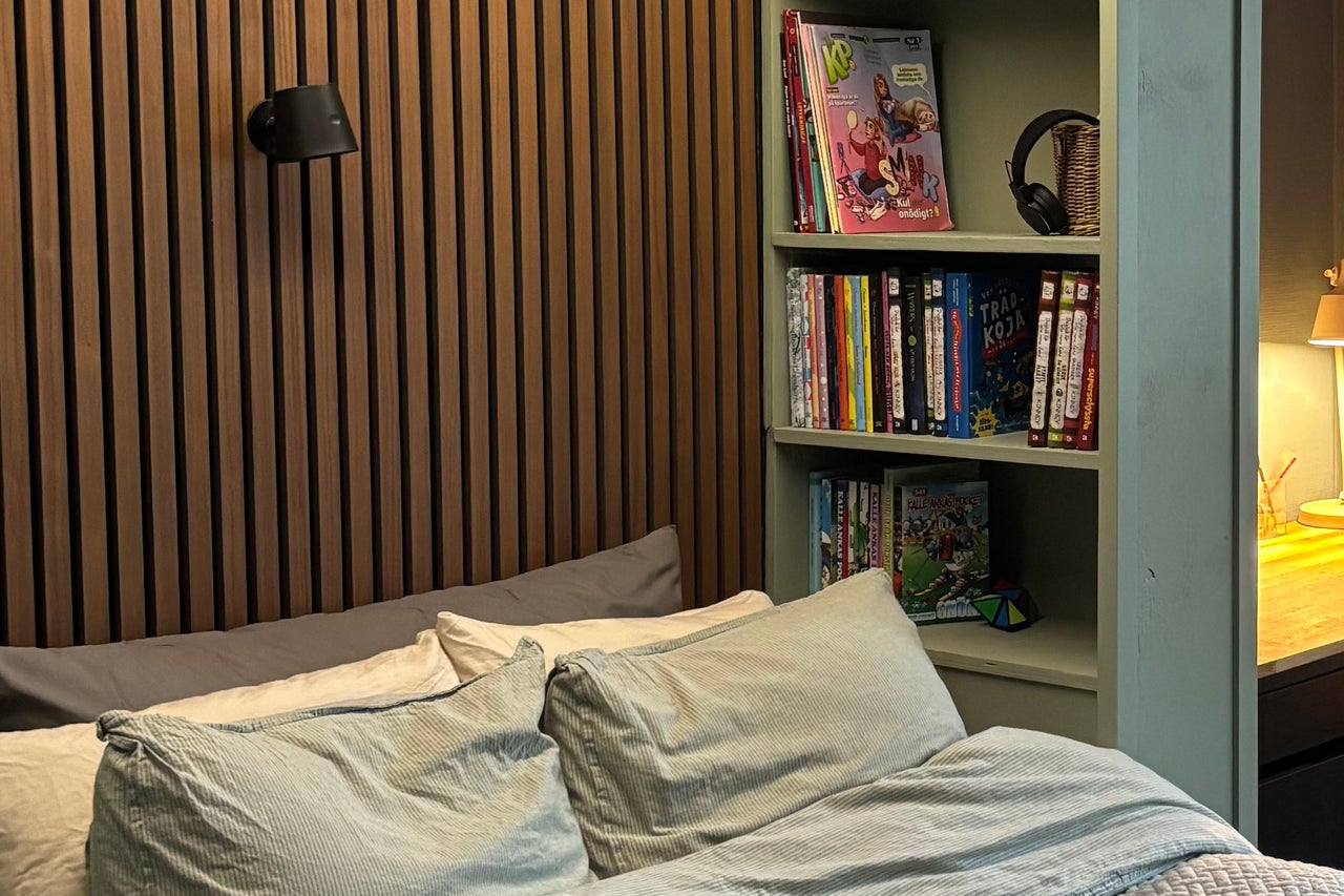 Sovrum med väggpanel av trälister, säng med randiga kuddar och en öppen hylla med böcker och en bordslampa.