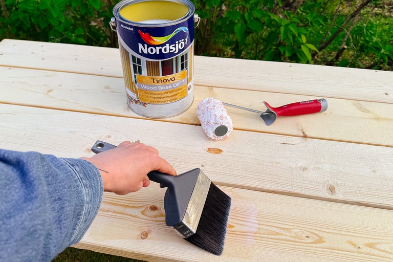 Träprimer appliceras med en pensel på en träyta, tillsammans med en färgroller och en färgburk.