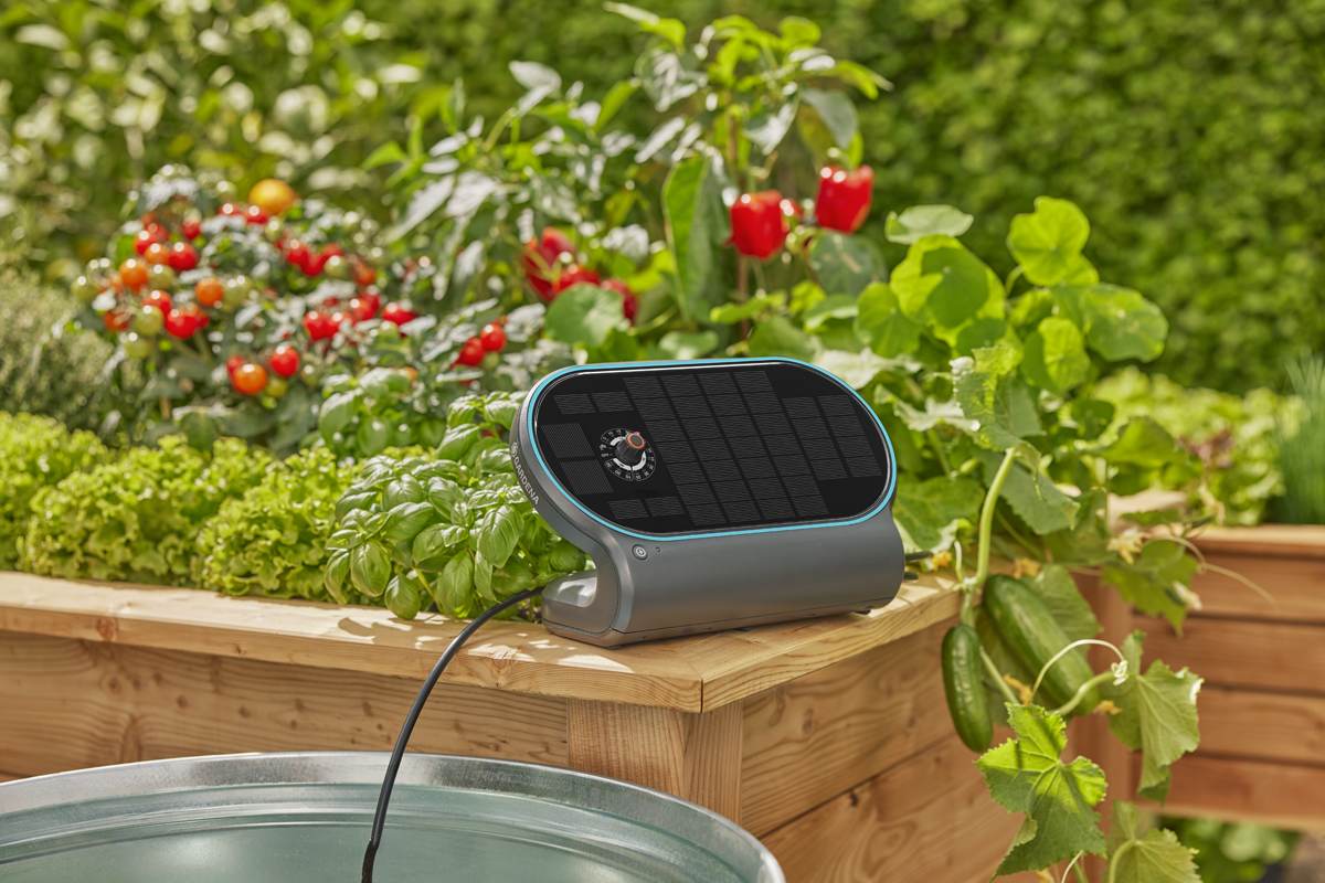 Gardena solcellsdriven bevattningsdator på en upphöjd odlingsbädd i trä bredvid en vattenbehållare i en trädgård.