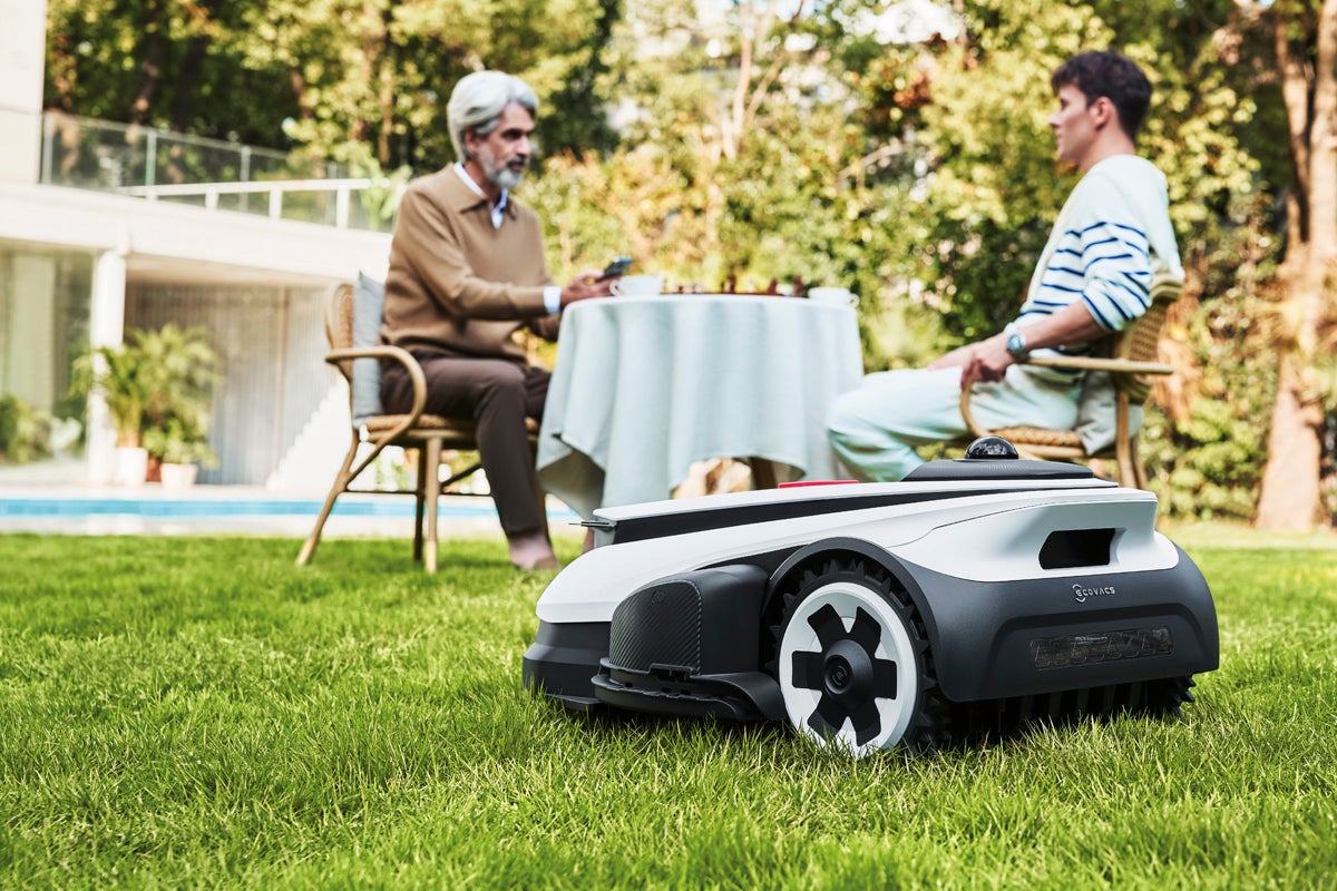 Robotgräsklippare på en grön gräsmatta i en välskött trädgård med pool och två personer vid ett bord i bakgrunden.