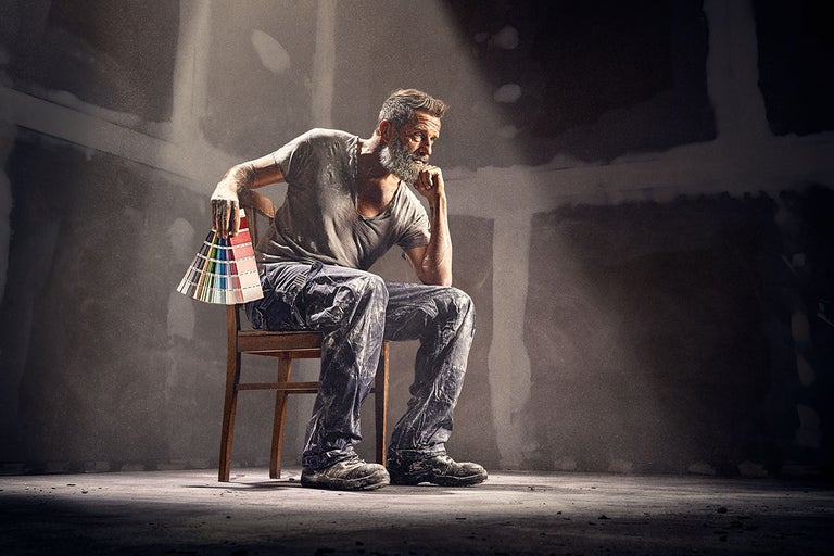 En man sitter på en stol och håller en färgfläkt i handen medan han funderar i ett rum med putsade väggar.