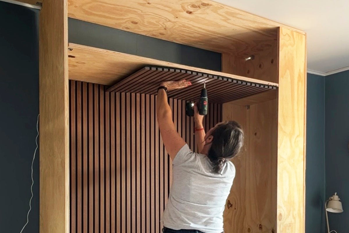 En person installerar trälister i taket på en platsbyggd hylla av plywood med hjälp av en batteridriven borrmaskin.