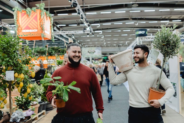 Två män bär växter och en blomkruka i ett ljust butiksutrymme under en Hornbach-logotyp