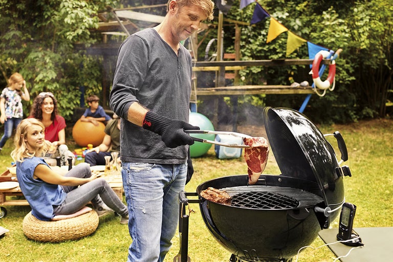 Man grillar kött på en klotgrill i trädgården, omgiven av familj och vänner.