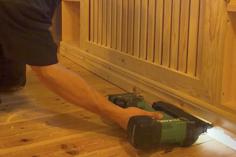 En person använder en Hikoki batteridriven spikpistol för att fästa en trägolvlist vid en elementbeklädnad i trä.