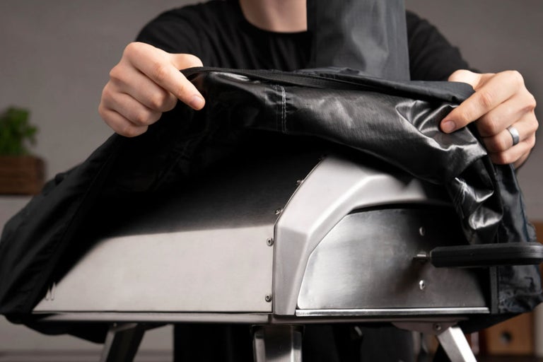 Person lägger ett svart skyddshölje över en pizzaugn i metall.