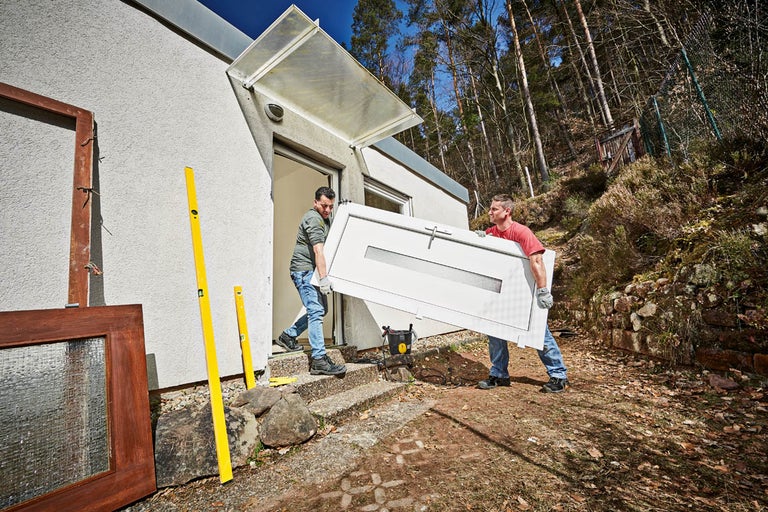 Två personer bär en ytterdörr för installation i en byggnad; vattenpass och en fönsterkarm syns i förgrunden.