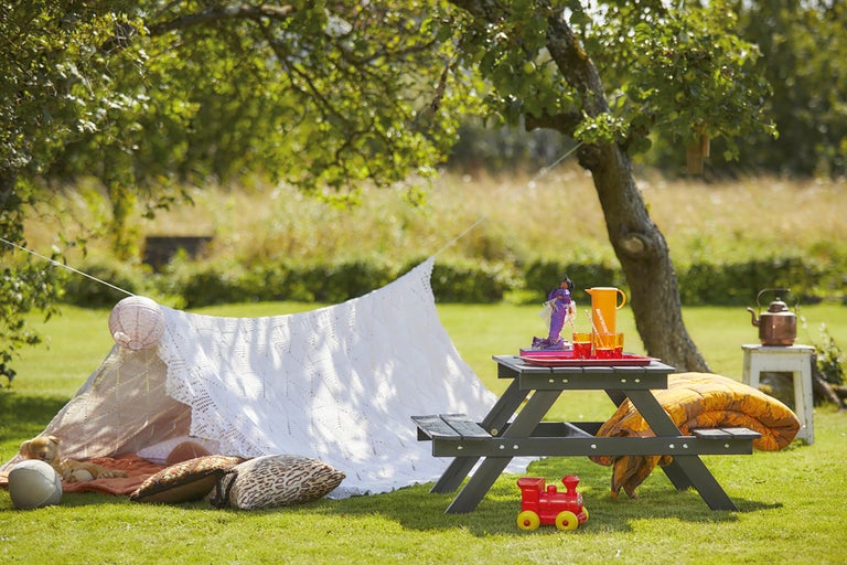 Trädgårdsscen med picknickbord, leksaker och ett improviserat tält under ett träd