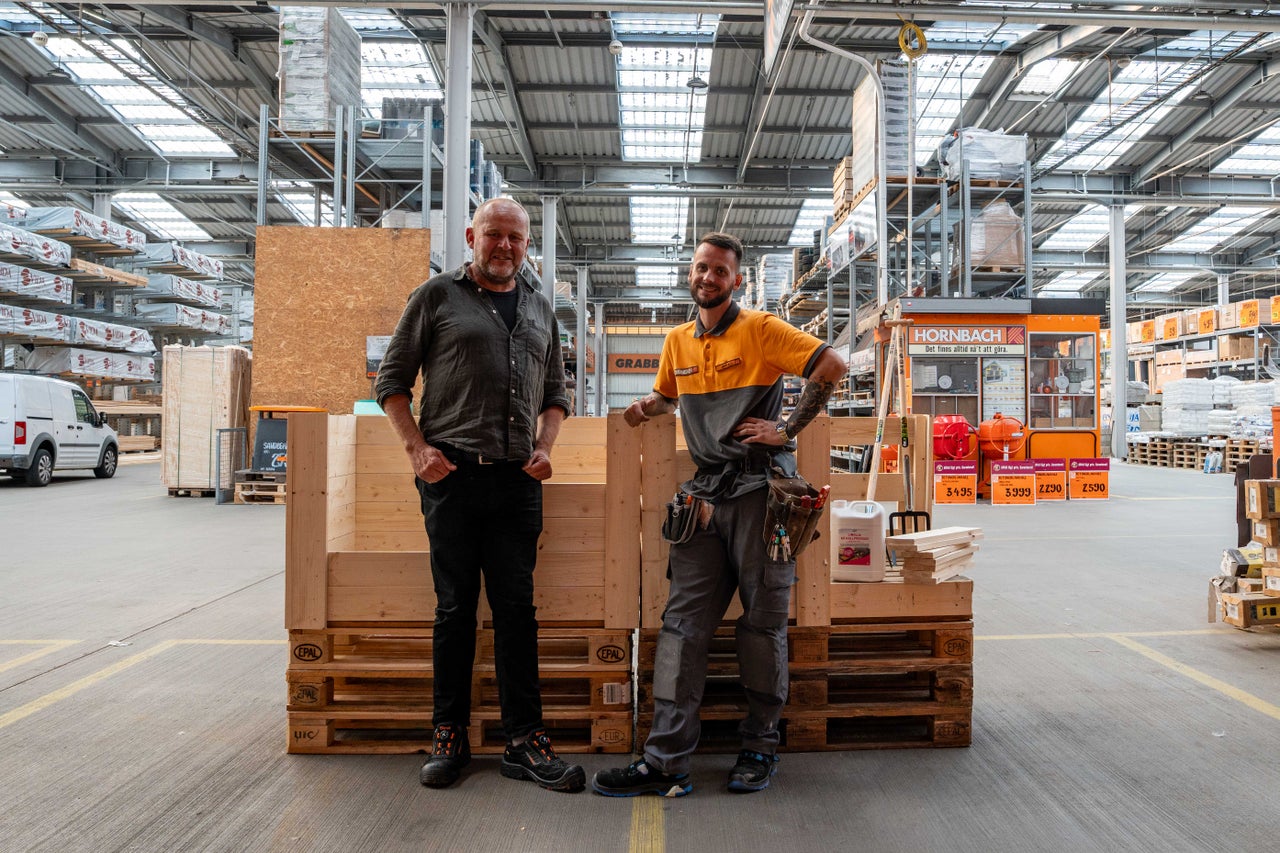 Två personer står i ett Hornbach byggvaruhus framför träkonstruktioner på europallar i en stor, välbelyst lagerhall.