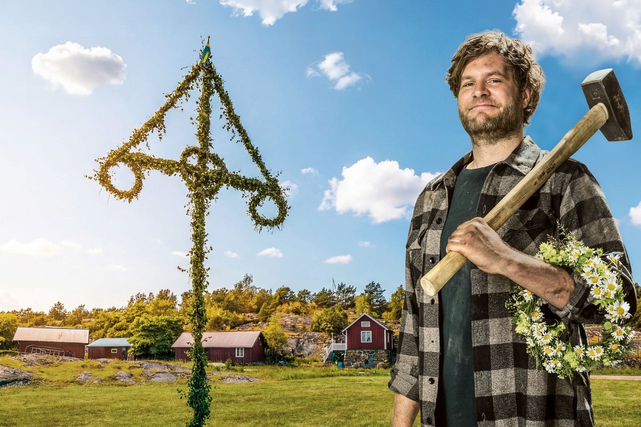 En man i rutig skjorta som håller en slägga och en blomsterkrans framför en midsommarstång och trähus i ett svenskt landskap.