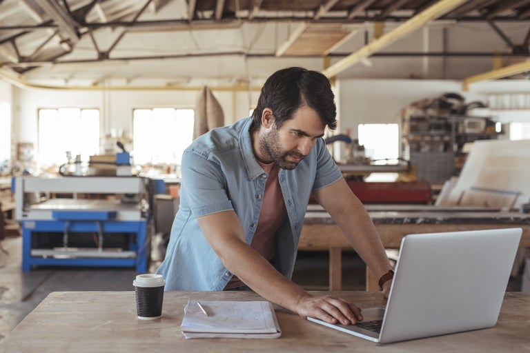Man arbetar på en bärbar dator i en verkstad, skisser och kaffe på bordet.