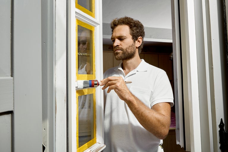 En man målar en fönsterkarm med en pensel, fönstret är maskerat med tejp.