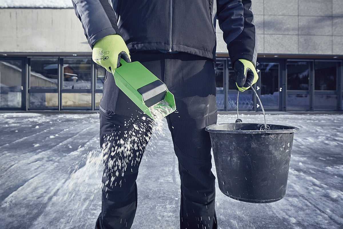 Man strör vägsalt med en saltspridare och en hink på en isig yta.