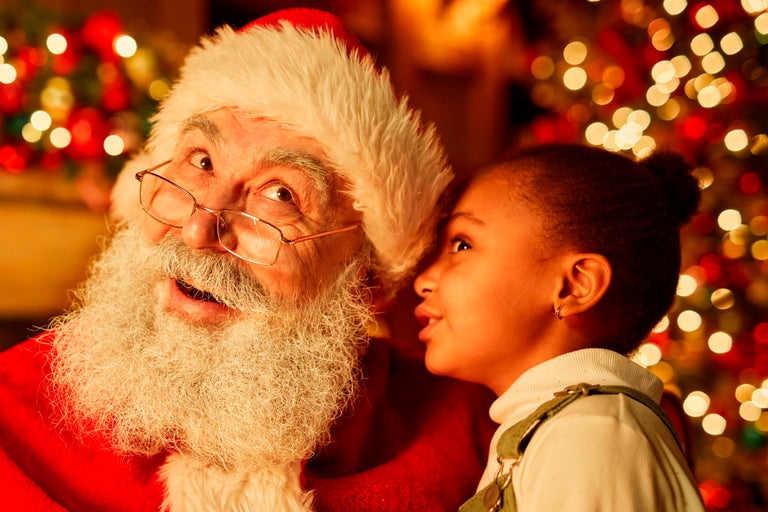 Närbild av Santa Claus med skägg och glasögon, medan ett barn viskar något i örat på honom.