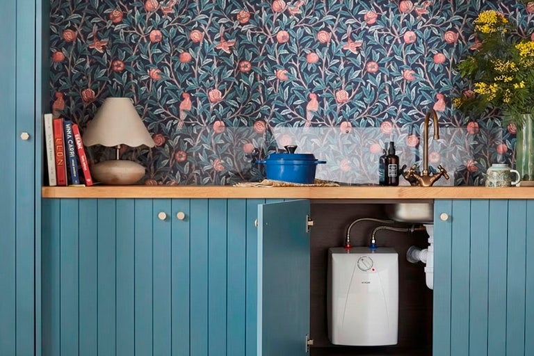 Köksscen med blått underskåp, diskho, kran, kastrull, varmvattenberedare och blommig tapet