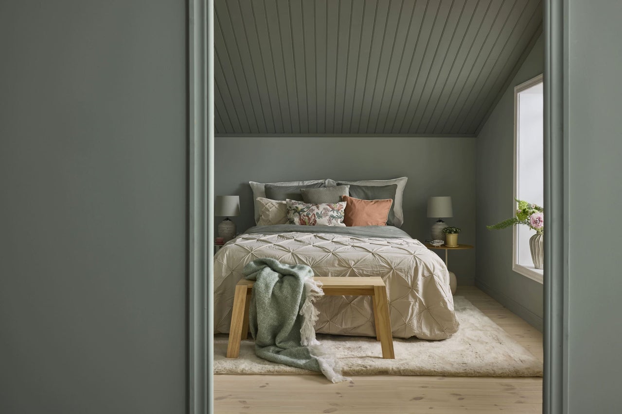 Mysigt sovrum med dovt gröna väggar, panelklätt tak, säng med quiltat täcke, träbänk och mjuk matta.