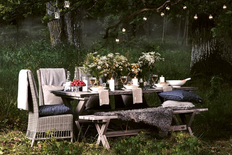 Atmosfärisk trädgårdsscen med matbord, bänk, stol, kuddar, porslin, blommor och utomhusbelysning