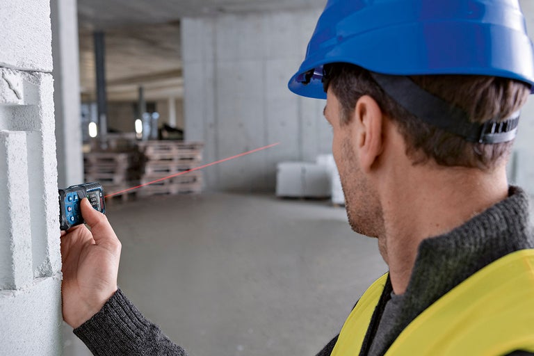 En byggnadsarbetare med hjälm använder en laseravståndsmätare på en byggarbetsplats.