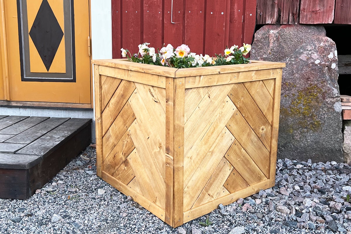 Kvadratisk blomlåda i trä med fiskbensmönster, planterad med penséer, placerad utomhus på grus.