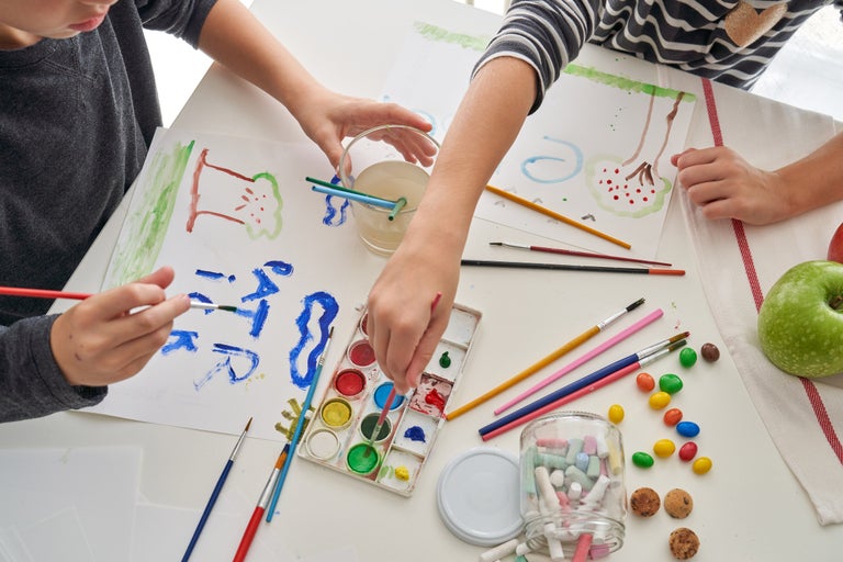 Scen med barn som målar med akvarellfärger, penslar, vattenkopp, krita och papper, arrangemang på ett bord.