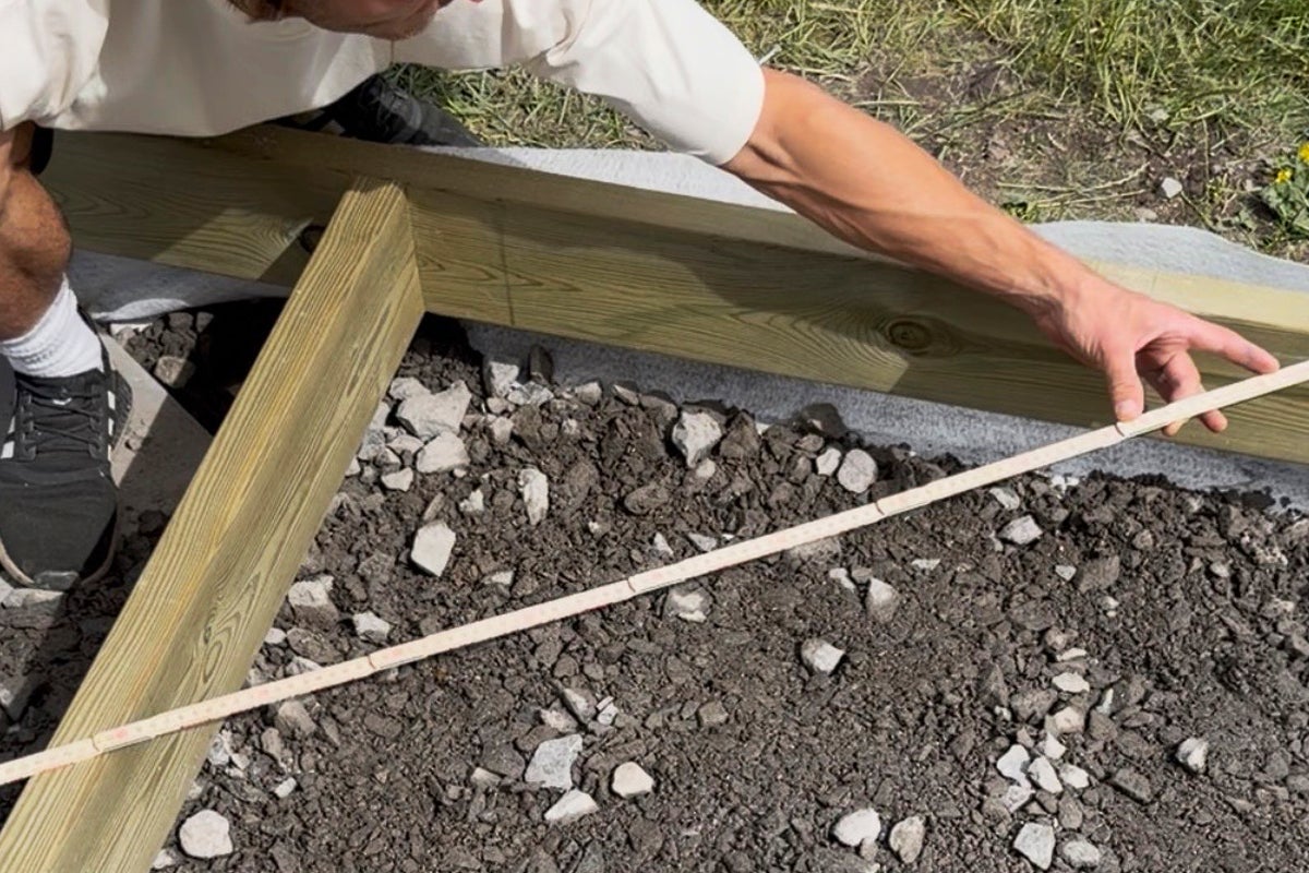 En person mäter en altans träram med en tumstock på ett underlag av grus och fiberduk.
