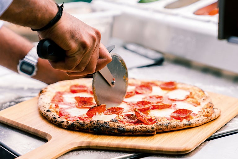 En person skär en salami pizza på en träbräda med en pizzaskärare.