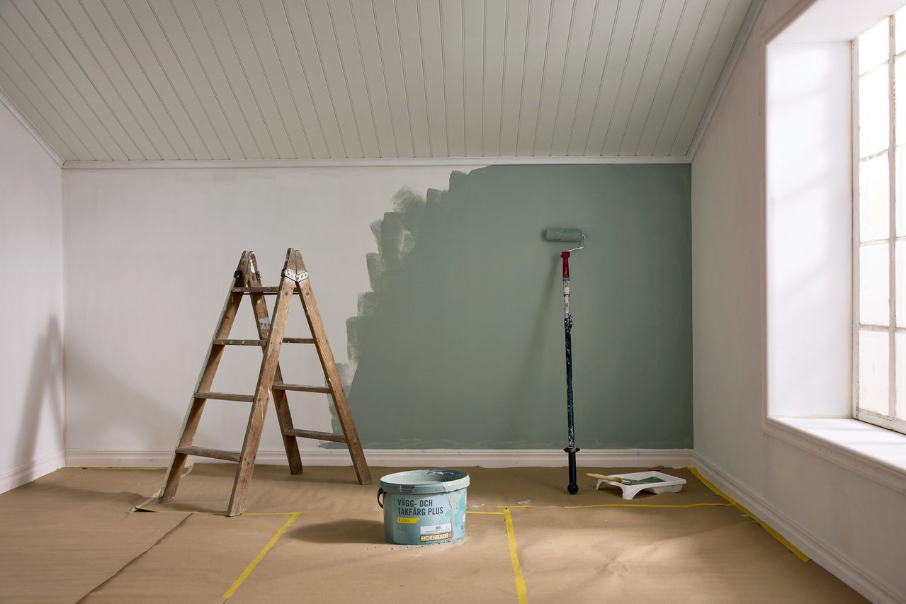 Ett ljust rum under renovering med en halvmålad grön vägg, trästege, Hornbach färg hink och målarverktyg på täckpapper.