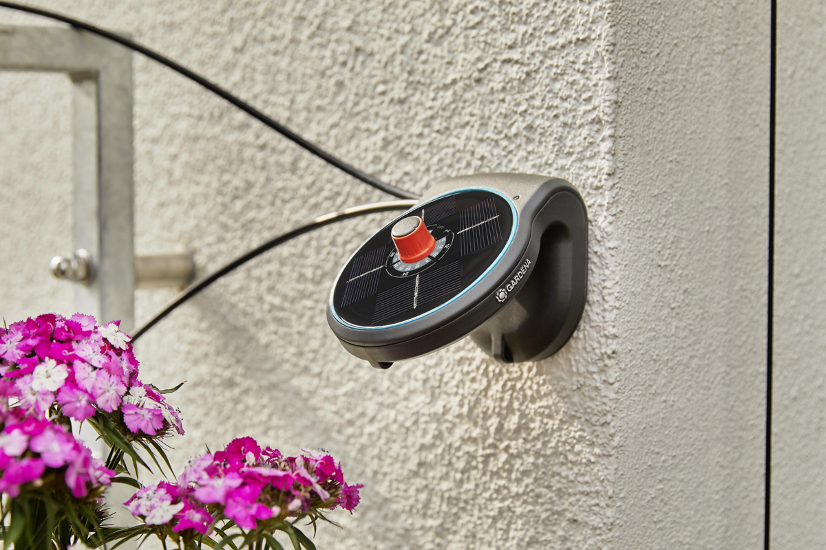 Gardena bevattningsdator med solpanel monterad på en vägg bredvid blommor