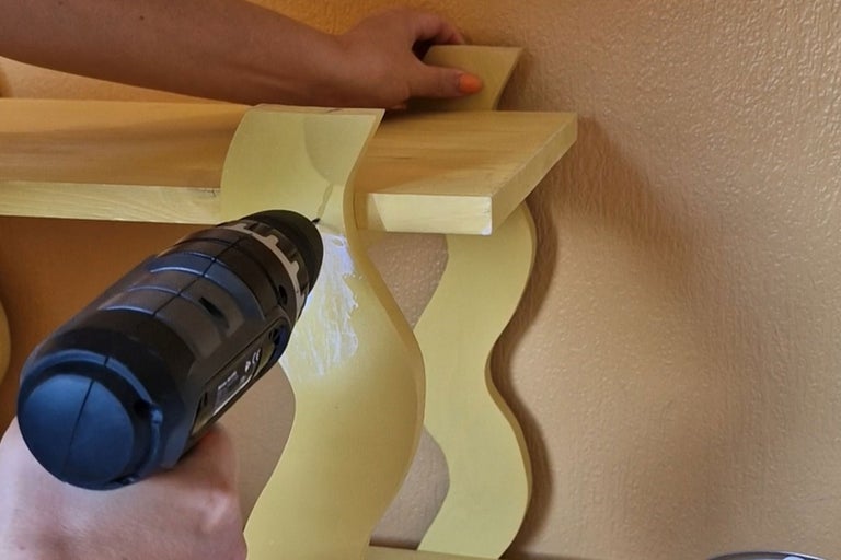 En person fäster en vägghylla i trä på väggen med en sladdlös borrmaskin.