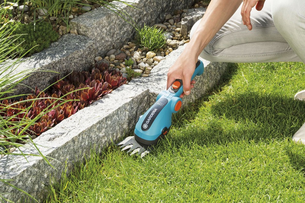 Person klipper gräs med en Gardena batteridriven grässax längs en stenmur