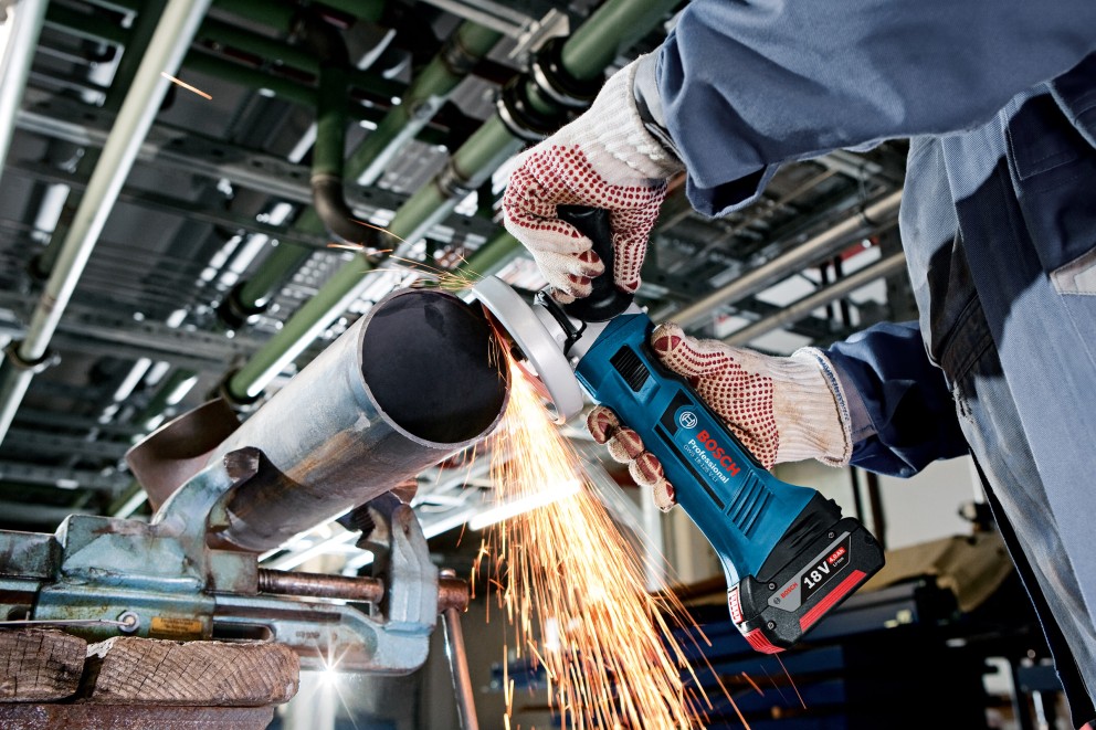 Batteridriven vinkelslip används för att slipa ett metallrör, fixerat i ett skruvstäd. Gnistor flyger. Bosch logotyp synlig.